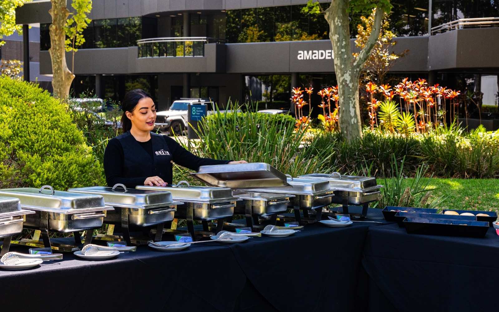 Person setting up a table with food containers outdoors, with 'Amade' building in the background.